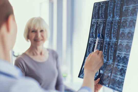 Focus on spine X-ray being held by specialist in clinic. She is pointing with pen at it and discussing with smiling mature lady who is standing beside. Copy space in left side