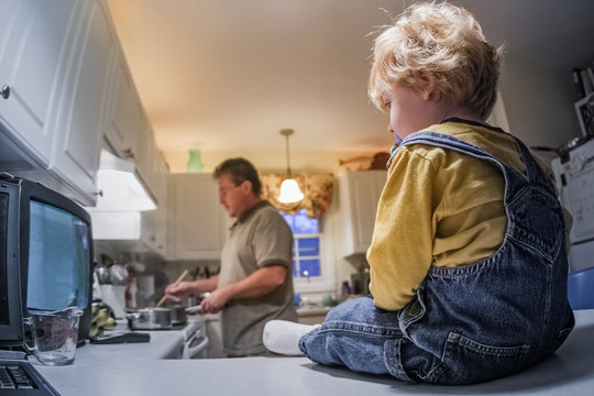 Toddler boy sitting on kitchen counter watching tv while father prepares dinner