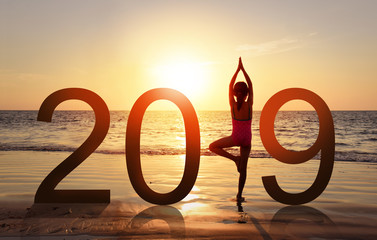 Happy new year card 2019. Silhouette of healthy girl doing Yoga vrikshasana tree pose on tropical beach with sunset sky background, watching the sunset, standing as a part of the Number 2019 sign.
