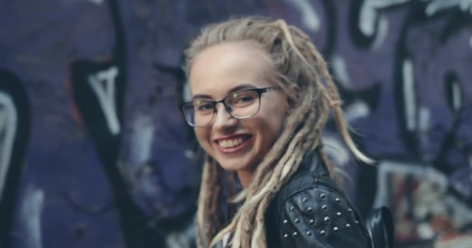 Rear of the close up of the hipster pretty young girl in glasses and with dreadlocks turning her head smiling cheerfully to the camera on the graffity background. Close up. Portrait. Outdoor.