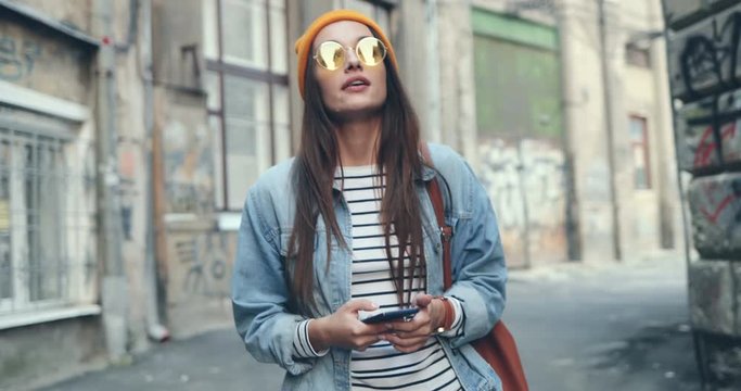 Caucasian joyful young beautiful woman in the hipster style walking the slum street and texting on the smartphone. Outdoor.