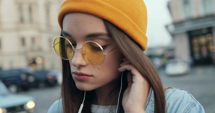 Close up of the young pretty girl in sunglasses and hat putting in ears headphones as listening to music on the street. Outdoor.