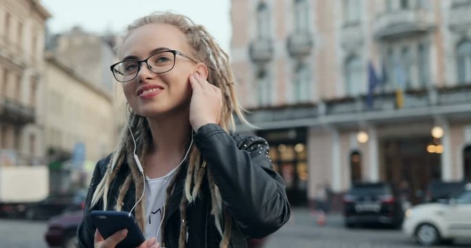 Portrait shot of the young pretty girl in glasses and with dreadlocks enjoying of listening to the music while standing in the city. Outside.