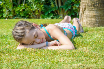Beautiful girl blindly sunbathes in the sun lying on green grass near palm tree.