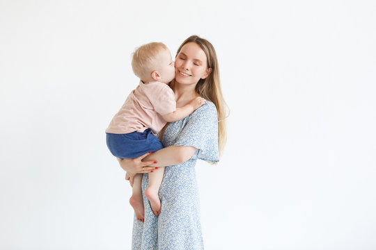 Family, Motherhood, Parenting, People And Child Care Concept. Happy Mom Embraces Adorable Baby Over White Wall Background. Little Cute Kid Kissed His Mother With Love. Family At Home Relaxing