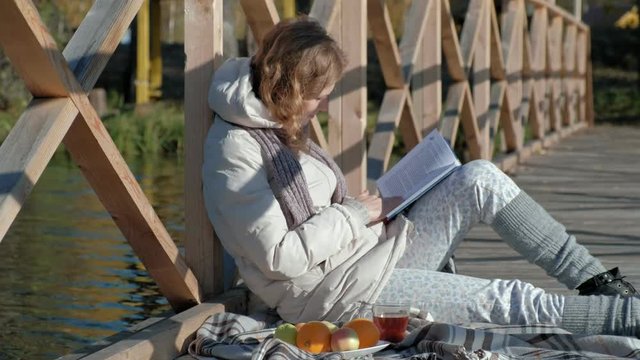 a woman on the bridge near the autumn river reads a book and drinks tea