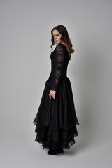 full length portrait of brunette girl wearing long black lace gown with corset.  standing pose with back to the camera, grey studio background.