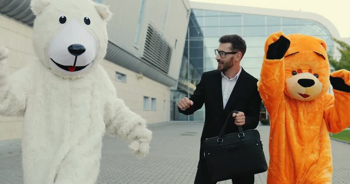 Funny picture of the cheerful Caucasian man in a suit and with a bag in a hand stepping the street and dancing while having fun with two big growth dolls of bears.