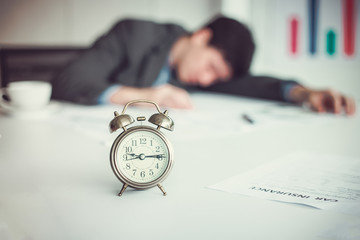 Blurred background of Exhausted businessman sleeping head on table with document in his office. Focus on a clock.