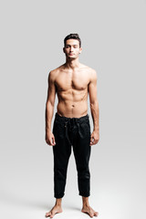 Fototapeta premium Dark-haired handsome young dancer with bare torso wearing a black sports pants is standing on a white background