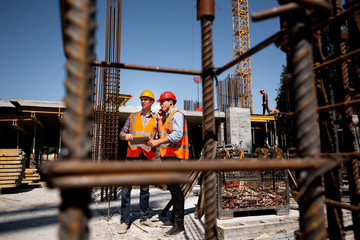 Architect  and structural engineer  in orange work vests and  helmets discuss a building project on the open air building site with a lot of steel frames