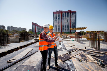 Architect  and construction manager dressed in orange work vests and helmets discuss documentation...