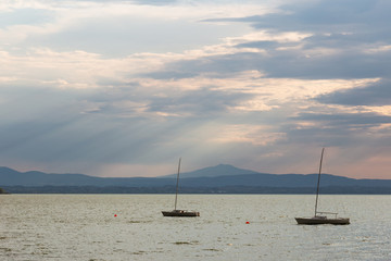 Fototapeta premium A couple of empty, little sailboat on a lake, beneath a moody sky with sun rays filtering through