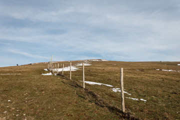 Wooden fence posts and barbed wire marking the property line of a prairie with dried grass and patches of snow under blue sky with white clouds