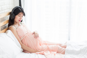 Relaxing Pregnancy woman  in pink dress lisening music from headphone on the bed in bedroom.
