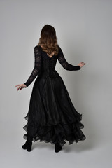 full length portrait of brunette girl wearing long black lace gown with corset.  standing pose with back to the camera, grey studio background.
