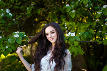 a young teenager girl with long dark hair in a white dress standing under a blooming apple tree