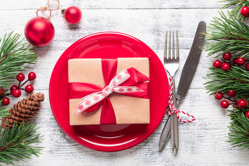 Christmas table setting with red plate, gift box and silverware on light wood background. Fir tree...