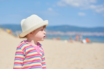 Little two year old girl with blue eyes in a bright wicker hat, standing on the beach near the sea