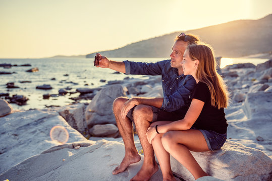 Father and daughter sitting on a rocky beach and taking selfie