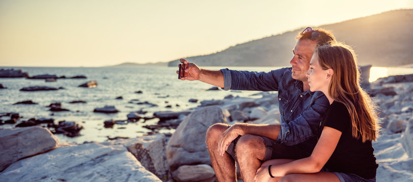 Father and daughter sitting on a rocky beach and taking selfie