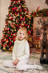 Christmas portrait of happy blonde child girl in white sweater siting on the floor near the Christmas tree and wooden toy horse. New Year Holidays