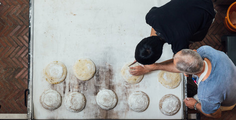 Artisan baker teaches his apprentice to slice yeast bread dough before bake in electric oven.
