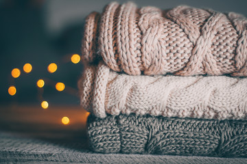 Stack of cozy knitted sweaters and garland lights on wooden background. Autumn-winter concept....