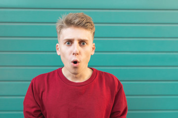 Portrait of a surprised young man looking at the camera at the background of a turquoise wall. Amazed teenager in a red sweatshirt on a green background.