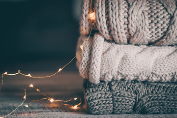 Stack of cozy knitted sweaters and garland lights on wooden background. Autumn-winter concept. Magic, cozy and mood time