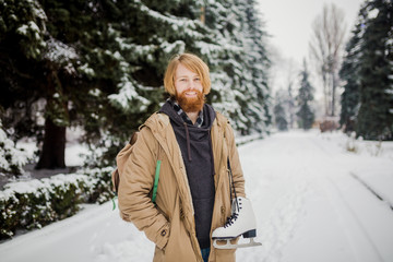 Winter sports theme. Portrait. Handsome young Caucasian guy with long hair and red beard. Model poses in winter snow park against background of the forest on the shoulder of a pair of leather skates