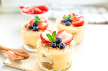 homemade, exquisite dessert tiramisu in glasses decorated with strawberry, blueberry, mint on white wooden table