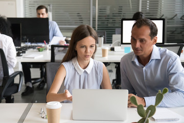 Focused office colleagues discussing online project looking at laptop screen, female employee explaining computer task to male coworker talking working together, help and mentoring teamwork concept