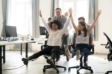 Diverse excited workers having fun good time together enjoy riding on chairs on friday, happy...