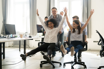 Diverse excited workers having fun good time together enjoy riding on chairs on friday, happy...