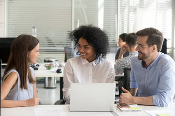 Three friendly diverse colleagues work together on laptop, smiling multiracial mentors training teaching intern with computer, multi-ethnic office coworkers team talking at meeting, teamwork concept