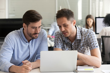 Focused serious male coworkers talking working together on computer project discussing software with laptop analyzing online marketing results in office, mentor teaching intern, cooperation concept