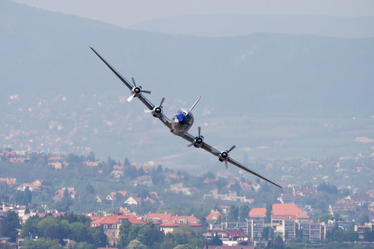 Vintage four-engine propeller airliner flying low over city buildings and mountains. Classic aviation concept.