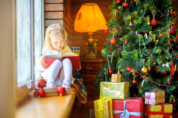 Fototapeta na wymiar blonde girl sits on the sideboard and reads Christmas tales, lots of gifts under tree, Christmas morning