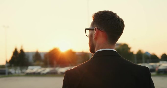 Rear Close Up Of The Caucasian Man In Glasses And Business Style Walking To The Parking Outdoor On The Sunset. Back View.