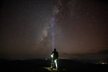 Silhouetted person stands atop boulder with light beam looking up at the milky way and sunrise.