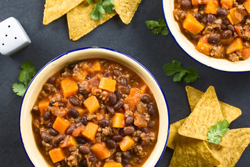 Homemade chili con carne with mincemeat, red and black beans, tomato sauce and pumpkin, tortilla chips on the side, photographed overhead on slate (Selective Focus on the chili)