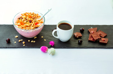 Berry yoghurt with granola in a glass bowl, cup of freshly brewed coffee and chocolate on black stone tray.  Served breakfast.