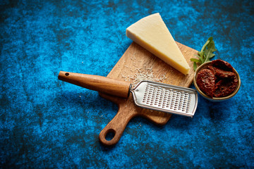 Grated parmesan cheese and metal classic grater placed on wooden cutting board. Dried tomatoes on side. Blue background. View from above.