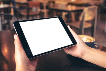 Mockup image of hands holding black tablet pc with blank white screen with coffee cup on wooden table in cafe