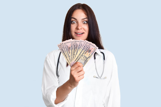 Shocked female doctor holds dollar bills, being rich, involved in corruption, cant believe she has much money, makes business on patients, poses against light blue background. Medicine is paid