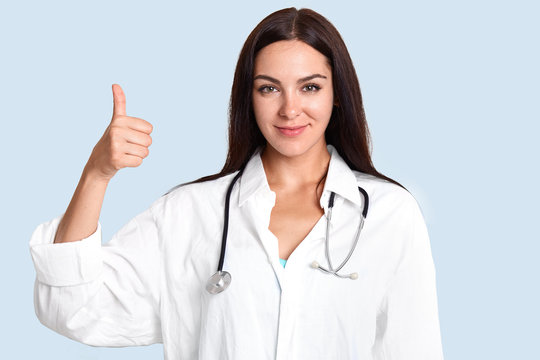 Horizontal shot of delighted female doctor keeps thumb raises, aprroves or gives agreement, wears white medical robe with stethoscope, isolated over blue background. Body language, medicine.