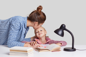Photo of young woman cares of her child, kisses daughter in forehead, praises her to study well,...