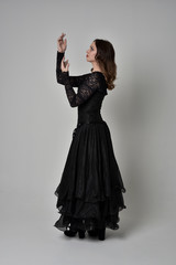 full length portrait of brunette girl wearing long black gown with corset. standing pose on grey studio background.