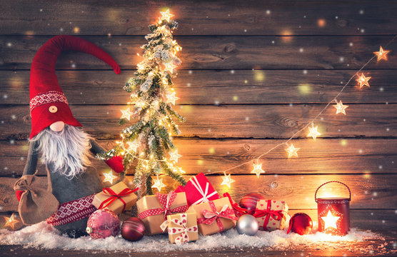 Santa Claus or dwarf holds a fir tree with Christmas lights surrounded by gift boxes and glowing lantern